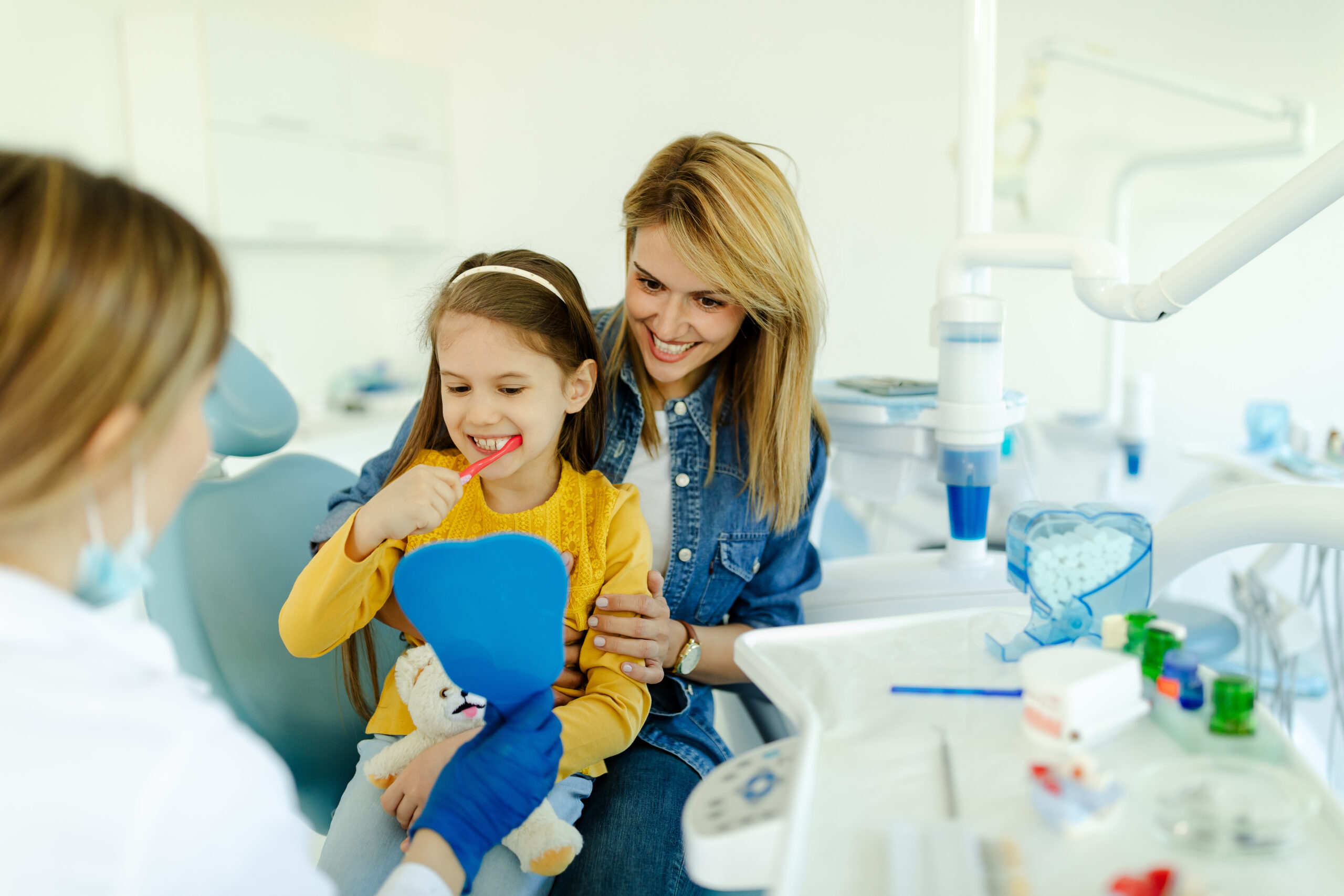 Girl looking in the mirror and brush her teeth after dental procedure while mother sitting near her for support.