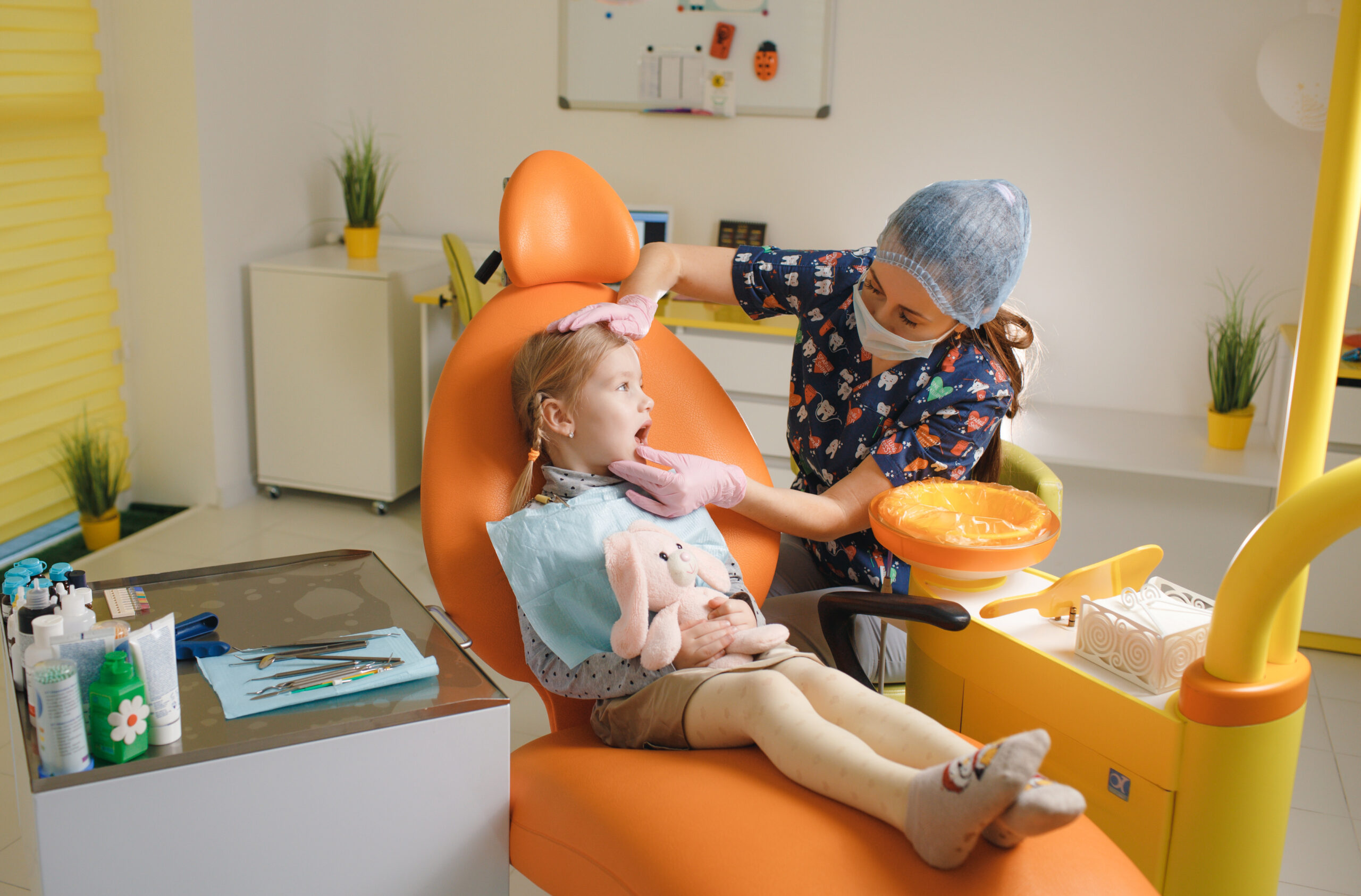 A little cute smiling girl is sitting in an orange dental chair showing a woman dentist a sore tooth.